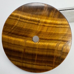 May include: A circular tiger's eye gemstone with a central hole. The stone displays bands of golden brown and yellow, forming a striped pattern. The surface is smooth and reflective, highlighting the natural variations. The stone is approximately 6 cm in diameter.