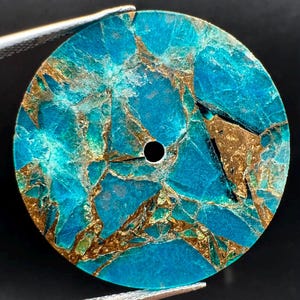May include: A circular watch dial featuring a vibrant turquoise blue surface with intricate, jagged gold-coloured metallic veining throughout. The dial has a single, centred circular hole for the watch hands and a smooth, flat finish.