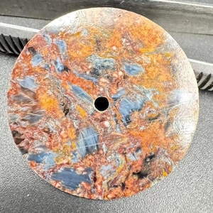 May include: A round, patterned stone with a central hole. The stone features a mix of colors, including orange, blue, and brown, creating a marbled effect. The surface appears polished and smooth, with a dark hole in the center.