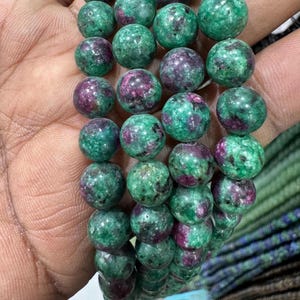 Peut inclure: Un collier de perles rondes et polies présentant un motif marbré de vert vif et de rose magenta profond. Chaque perle affiche une surface lisse et brillante avec des variations de couleur naturelles et des inclusions sombres.