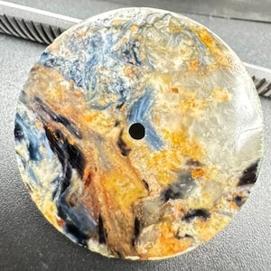 May include: A round, polished gemstone with a drilled hole. The stone features a swirling pattern of blue, yellow, orange, and black hues. The gemstone's surface has a glossy finish, highlighting the natural color variations.