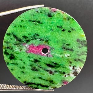 Puede incluir: Un colgante de piedra preciosa circular y plano que presenta una base de color verde vibrante con inclusiones dispersas de color verde oscuro y negro, acentuado por una mancha magenta rosada irregular cerca del centro con un pequeño orificio redondo.