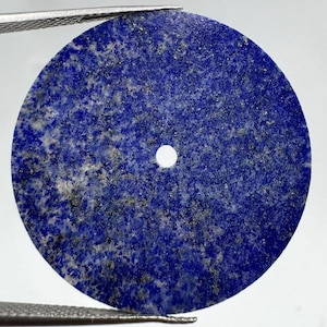 Puede incluir: Un colgante circular de lapislázuli con un color base azul profundo, inclusiones naturales de calcita blanca y motas de pirita dorada metálica. El disco de piedra plano tiene un pequeño orificio circular centrado para su sujeción.