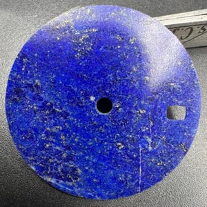 May include: A circular watch face crafted from lapis lazuli, a deep blue stone with gold inclusions. It has a small circular hole and a square aperture, probably for the watch hands and stem. The diameter is approximately 3.5 cm.