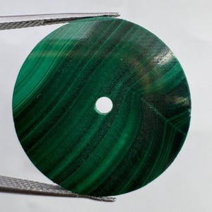 Pode incluir: Um disco circular de malaquita com faixas verde-escuras e padrões concêntricos com um pequeno orifício circular centralizado. A pedra apresenta uma superfície lisa e polida com variações naturais na intensidade da cor em toda a forma.