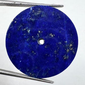 Könnte beinhalten: Ein runder, tiefblauer Lapislazuli-Edelstein mit einem kleinen Loch in der Mitte. Der Stein hat natürliche Goldflecken und wird von einer Pinzette gehalten. Der Edelstein hat eine satte, gesättigte Farbe.