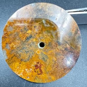 May include: A round, polished stone with a central hole. The stone features a blend of earthy colors, including shades of brown, yellow, and black, creating a natural, textured appearance. The stone's surface is smooth and reflective.