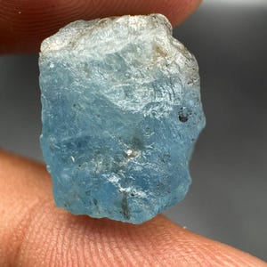 May include: A raw, unpolished aquamarine gemstone with a translucent light blue color. The stone features a natural, irregular crystalline structure with visible facets, internal inclusions, and a rough, textured surface throughout its body.