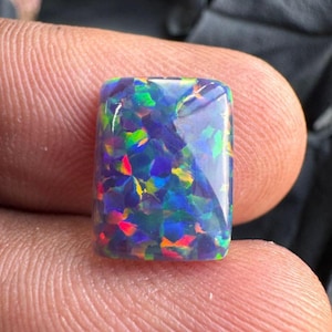 Puede incluir: Una piedra preciosa de ópalo negro rectangular con destellos de color vibrantes, incluyendo rojo, verde y azul. La superficie pulida refleja la luz, mostrando el juego de color interno de la piedra.