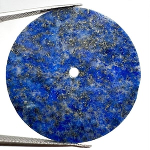 Può includere: Un ciondolo circolare in lapislazzuli caratterizzato da un colore di base blu intenso con inclusioni di pirite color oro sparse e venature minerali bianche. Il disco di pietra piatto presenta un piccolo foro circolare centrale per l'inserimento.