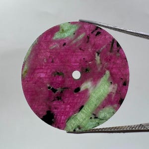 May include: A circular, flat disc of ruby zoisite featuring a vibrant mix of deep magenta-pink and pale lime-green mineral inclusions. The stone has a small, centred circular hole and is held by metal tweezers against a plain white background.