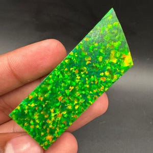 May include: A rectangular, polished gemstone with a vibrant green base color, speckled with yellow and orange inclusions. The stone is held in a hand, showcasing its unique color and texture. The gemstone's surface is smooth and reflective.