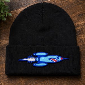 May include: A black knit beanie featuring an embroidered patch of a light blue rocket ship. The rocket has a pointed red nose, a shark-like mouth with white teeth, and a white exhaust trail extending from the back, all outlined in dark blue.