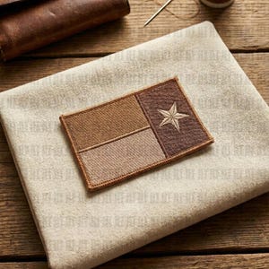 May include: A rectangular patch featuring the Texas state flag design. The patch is divided into three sections of varying shades of brown, with a white star in the rightmost section. The patch is placed on a beige fabric, with a wooden background.