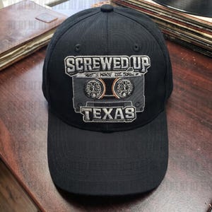 May include: A black baseball cap featuring an embroidered graphic of a cassette tape. The text reads SCREWED UP at the top and TEXAS at the bottom, with REST IN PEACE DJ SCREW written in smaller letters across the center of the cassette tape design.