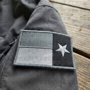 May include: A gray patch featuring the Texas flag design, with a white star on a black background and gray and light gray stripes. The patch is sewn onto a gray fabric.