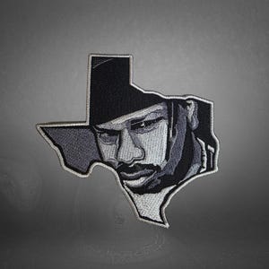 May include: Embroidered patch in the shape of Texas, featuring a grayscale portrait of a person wearing a hat. The patch has a white border and is likely designed to be ironed or sewn onto fabric.