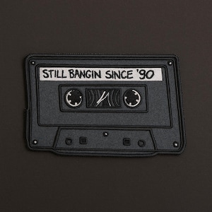 May include: Black cassette tape patch with the text "STILL BANGIN SINCE '90" in white. The patch has a detailed design with circular and rectangular features, and a central play symbol.