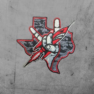May include: Embroidered patch featuring the outline of Texas in black and gray camouflage. A hand gesture with a red and white shark-shaped design is in the center. The patch has a red border.