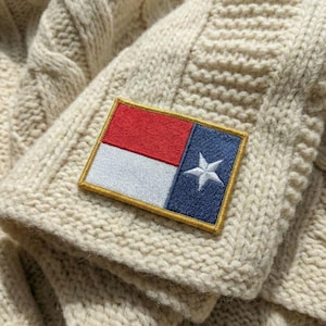 May include: Close-up of a cream-colored cable-knit sweater with a rectangular patch. The patch features a red and white section, a blue section with a white star, and a gold border. The sweater's texture is visible.