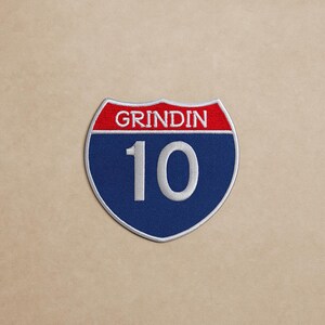 May include: Embroidered patch in the shape of a road sign. The patch features a red top section with the word "GRINDIN" in white, and a blue bottom section with the number "10" in white. The patch has a white border.
