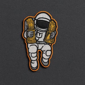 May include: Embroidered patch featuring a cartoon astronaut in a white spacesuit with gold accents, holding two bundles of gold bars. The patch has an orange border and is set against a dark gray background.