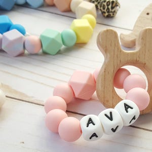 May include: A wooden bunny-shaped teether with a pink bead bracelet spelling "AVA". Other colorful silicone bead bracelets and a wooden elephant teether are also visible. The items are on a light wood surface.