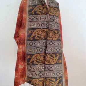 May include: A long, rectangular scarf with a paisley and geometric pattern in shades of orange, brown, and gold. The scarf is made of silk and has a fringed edge.