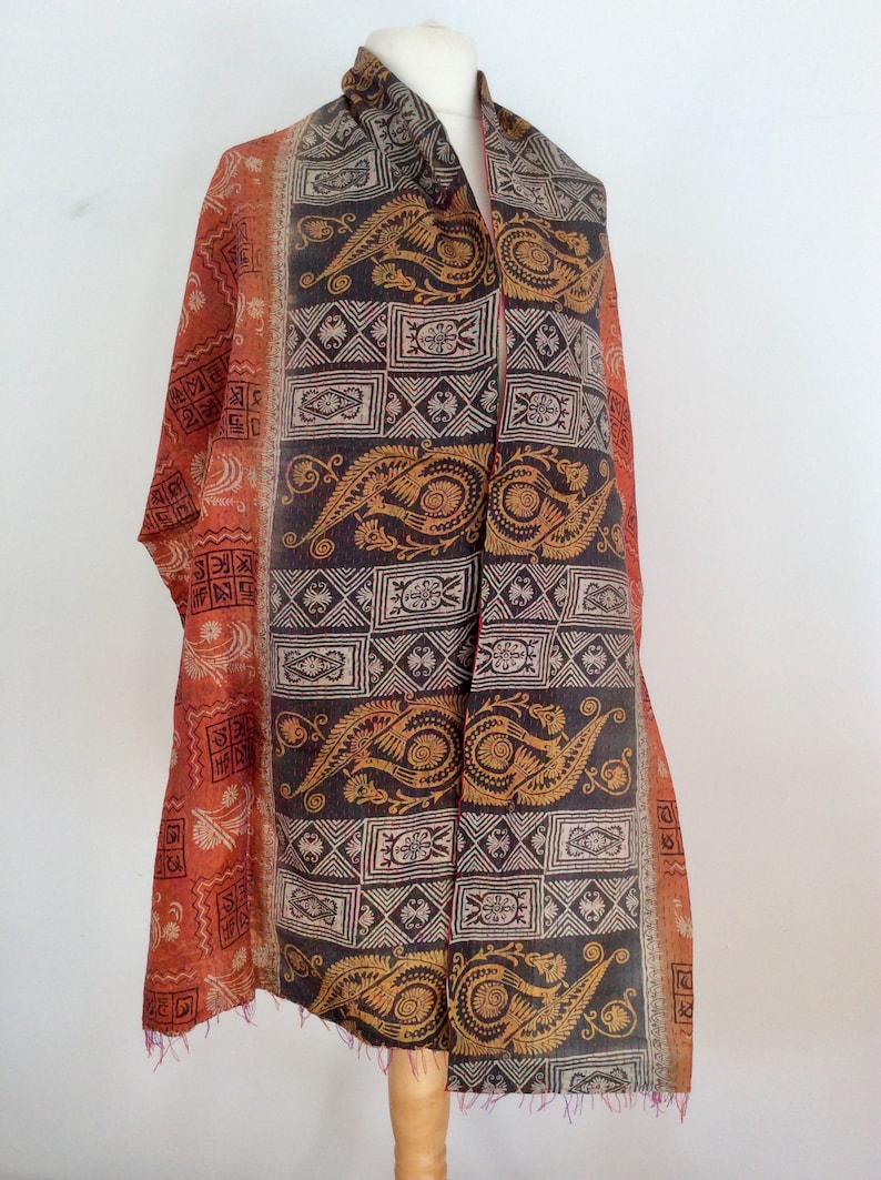 May include: A long, rectangular scarf with a paisley and geometric pattern in shades of orange, brown, and gold. The scarf is made of silk and has a fringed edge.