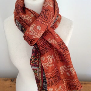 May include: A rust-colored silk scarf with a paisley pattern and a geometric design. The scarf is tied around a white mannequin's neck.