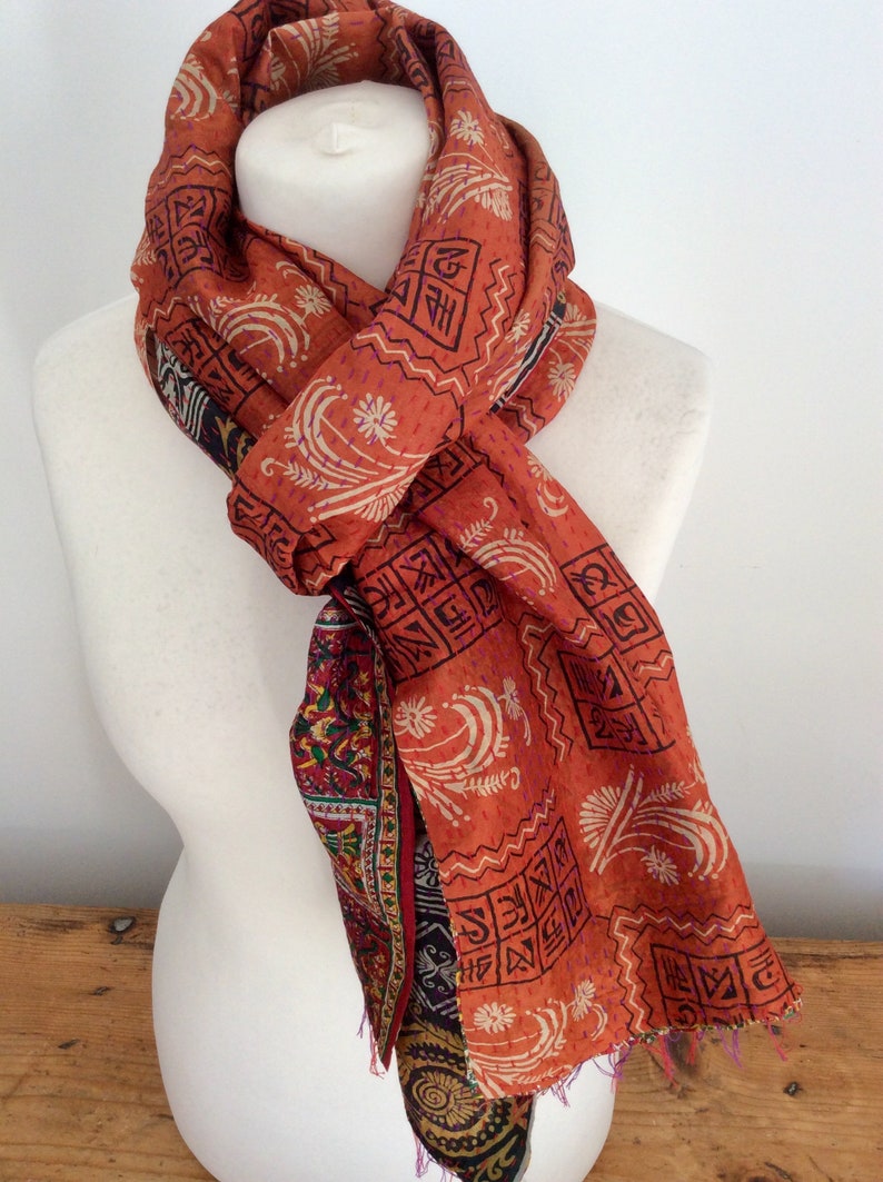 May include: A rust-colored silk scarf with a paisley pattern and a geometric design. The scarf is tied around a white mannequin's neck.
