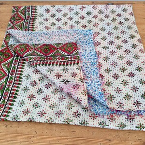 May include: A white kantha quilt with a red, green, and blue floral pattern. The quilt is folded in half and has a frayed edge.