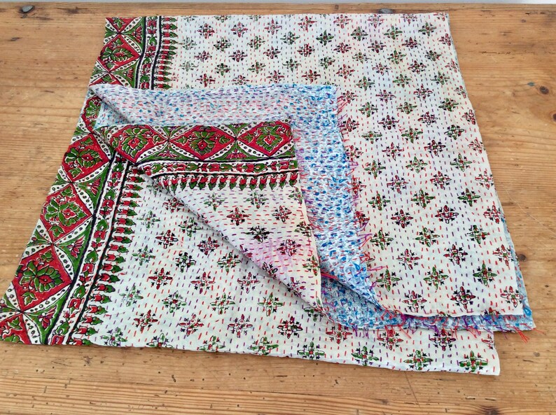 May include: A white kantha quilt with a red, green, and blue floral pattern. The quilt is folded in half and has a frayed edge.