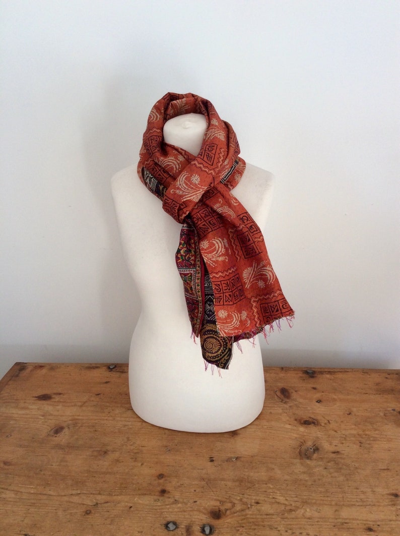 May include: A rust-colored silk scarf with a geometric pattern in black and white. The scarf is tied around a white mannequin's neck.