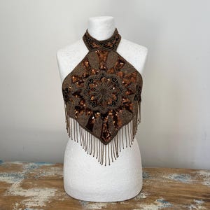 Puede incluir: Top halter marrón con intrincados abalorios y lentejuelas. El top presenta un cuello alto y un diseño floral central, con flecos de cuentas en el borde inferior. Se muestra en un maniquí blanco.