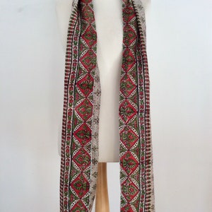 May include: A long, white scarf with a red, green, and black patterned border. The scarf is made of a soft, lightweight fabric and is perfect for adding a touch of color to any outfit.