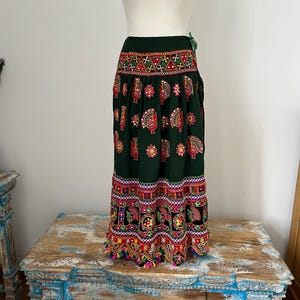Op de afbeelding: Een donkergroene rok met kleurrijke geborduurde patronen. De rok heeft een herhalend ontwerp van pauwen en bloemmotieven in rood, geel en blauw. De zoom is afgezet met een franje van kleurrijke kwastjes. De rok wordt op een mannequin getoond.