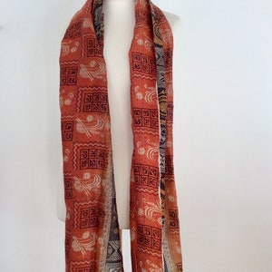 May include: A long, red and orange silk scarf with a black and white geometric pattern. The scarf is folded in half and draped over a white mannequin.