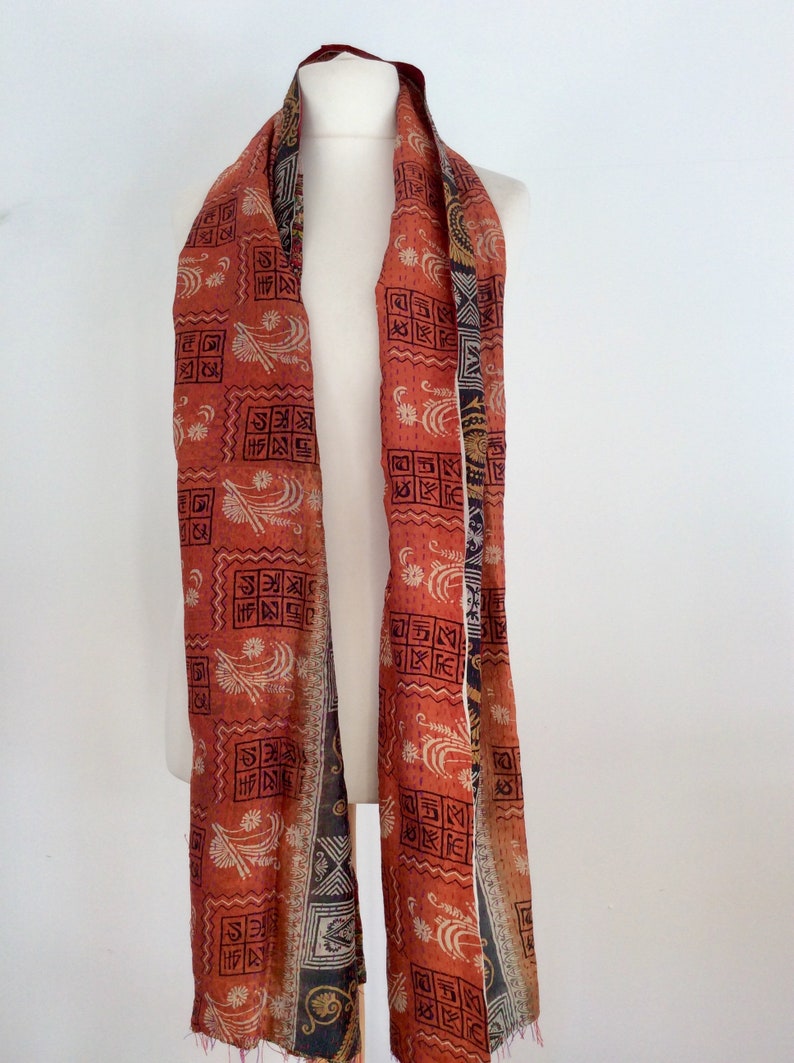 May include: A long, red and orange silk scarf with a black and white geometric pattern. The scarf is folded in half and draped over a white mannequin.