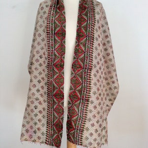 May include: A white cotton scarf with a red, green, and black floral print. The scarf has a fringed edge and is displayed on a wooden mannequin.