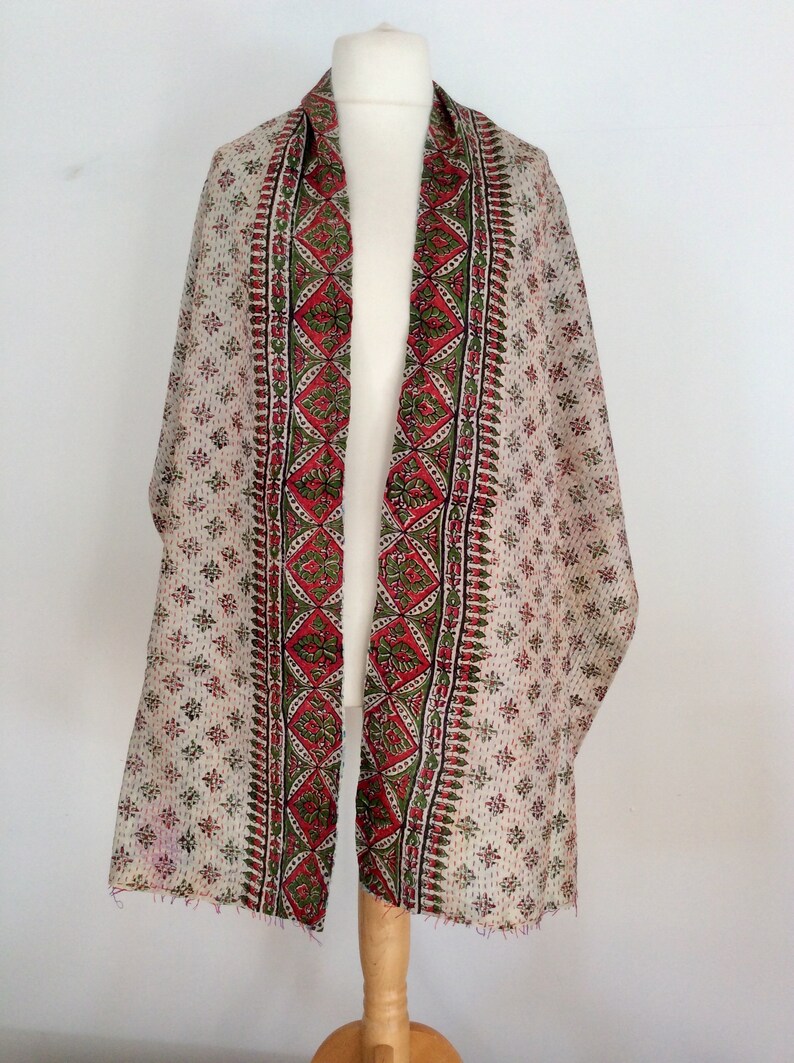 May include: A white cotton scarf with a red, green, and black floral print. The scarf has a fringed edge and is displayed on a wooden mannequin.