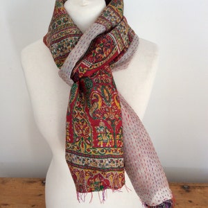 May include: A red, green, and yellow patterned silk scarf with a white and pink dotted border. The scarf is tied around a white mannequin's neck.