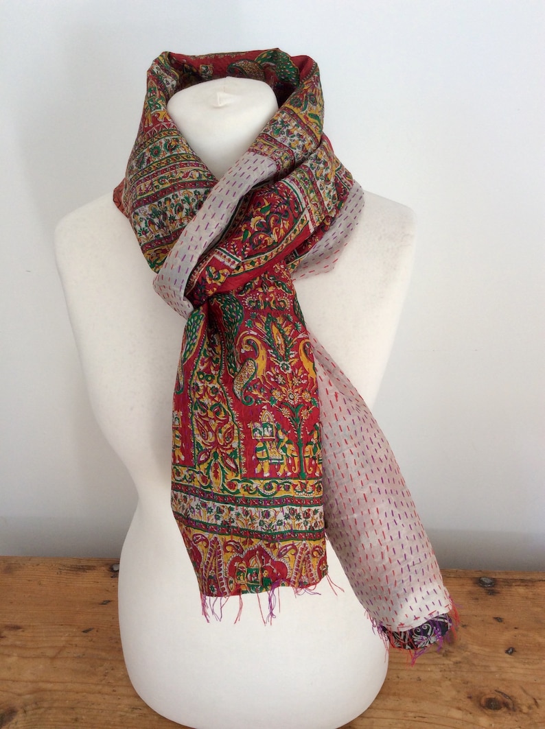 May include: A red, green, and yellow patterned silk scarf with a white and pink dotted border. The scarf is tied around a white mannequin's neck.