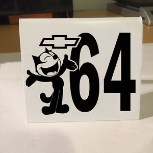 May include: A white sign with a black cartoon cat, a black Chevrolet logo, and the number 64.