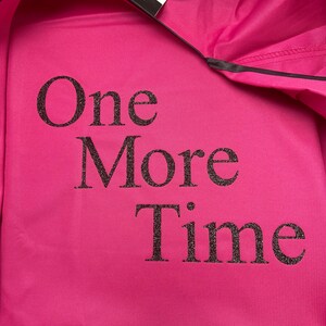 Op de afbeelding: Een felroze sweatshirt met de tekst "One More Time" in glinsterende zwarte letters. Het sweatshirt heeft een zilveren reflecterende streep aan de onderkant en een zwarte bies. Een label is zichtbaar.