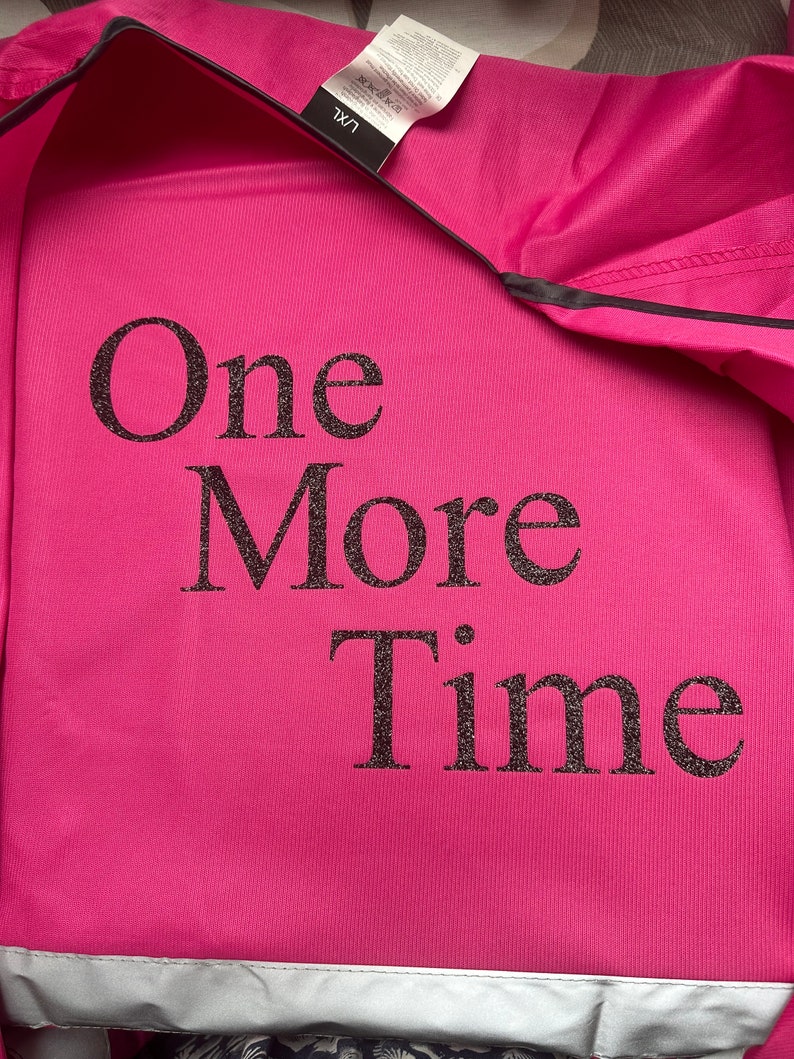 Op de afbeelding: Een felroze sweatshirt met de tekst "One More Time" in glinsterende zwarte letters. Het sweatshirt heeft een zilveren reflecterende streep aan de onderkant en een zwarte bies. Een label is zichtbaar.