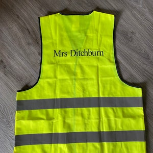 Op de afbeelding: Een felgele veiligheidsvest met reflecterende grijze strepen. De vest heeft de naam "Mrs Ditchburn" in zwarte letters. Ontworpen om de zichtbaarheid te vergroten, geschikt voor diverse veiligheidstoepassingen.