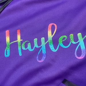 Op de afbeelding: Close-up van een paarse stof met de naam "Hayley" in een regenboogglitterlettertype. De stof heeft een getextureerd uiterlijk en de naam is in een cursieve stijl geschreven. De algehele esthetiek is levendig en gepersonaliseerd.