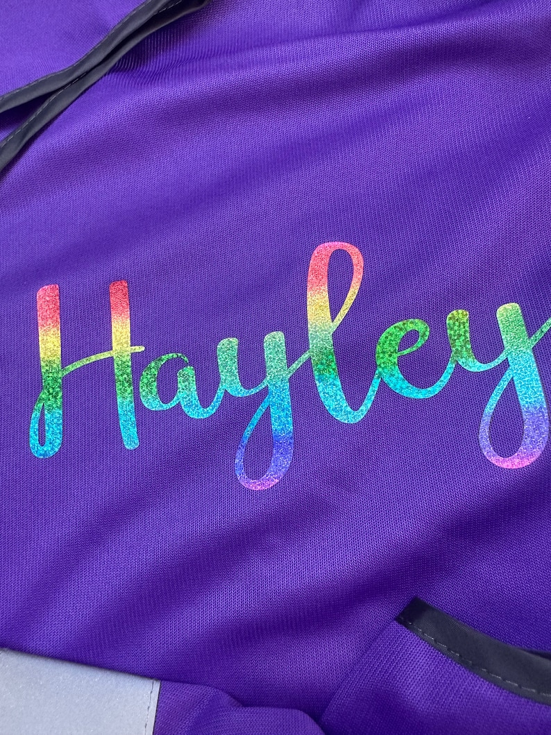 Op de afbeelding: Close-up van een paarse stof met de naam "Hayley" in een regenboogglitterlettertype. De stof heeft een getextureerd uiterlijk en de naam is in een cursieve stijl geschreven. De algehele esthetiek is levendig en gepersonaliseerd.