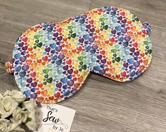 May include: A white sleep mask with a rainbow pattern of watercolor hearts. The mask has a tie on each side. The text "That's Sew by Jo" is on a white tag.